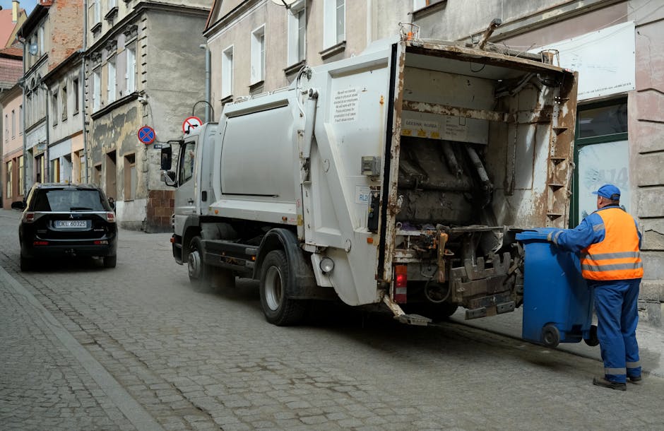 A street scene featuring a white waste collection truck operated by Rubbish Removal Kingston, positioned alongside a narrow urban road with cobblestone paving. The truck's rear load container is open, revealing rusted metal components and a hydraulic mechanism used for collecting rubbish. A worker dressed in a high-visibility orange vest and blue uniform, with protective gloves and a cap, is placing a blue wheeled bin into the truck’s designated opening. To the left, a dark-colored hatchback car is parked adjacent to the sidewalk, which is lined with historic-style buildings that have a weathered appearance, with some areas showing peeling paint and exposed brickwork. Street signs indicating parking restrictions are mounted on the building facades. The scene is lit by natural daylight, with muted shades enhancing the industrial and urban atmosphere, reflecting typical private waste collection activities carried out in residential or commercial areas, supporting the concept of alternative rubbish removal services outside of local authority collection.