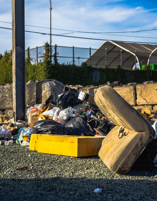 A pile of mixed rubbish including black plastic bags, an old beige car seat with visible fabric upholstery, and a yellow plastic container, all accumulated on a gravel surface next to a low stone wall. The background features a tall metal pole with electrical wires, a hedge, and a large outdoor tent structure with a curved roof, set against a partly cloudy sky. The scene is outdoors, possibly on a driveway or service area, illustrating an example of unmanaged waste or debris awaiting collection. The presence of diverse materials and the casual arrangement reflect a typical scenario where independent or scheduled rubbish removal services, such as those provided by Rubbish Removal Kingston, might be needed to clear the site effectively and responsibly.