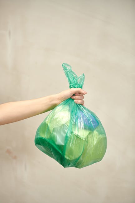 A person's arm is extended into the frame, holding a transparent green plastic bag filled with various waste materials, including light-colored and darker blue items visible through the bag. The bag is knotted at the top, with the excess plastic twisted into a small tail. The background is a plain, light-colored wall with a smooth surface, providing no additional environmental context. The scene appears to be indoors, possibly in a waste management or disposal setting, with natural or neutral lighting illuminating the scene evenly. The image visually represents waste collection or rubbish handling, relevant to independent or alternative rubbish removal services offered by companies like Rubbish Removal Kingston. The contents and handling of the bag reflect typical waste disposal practices, aligning with the context of private rubbish collection and environmental responsibility.