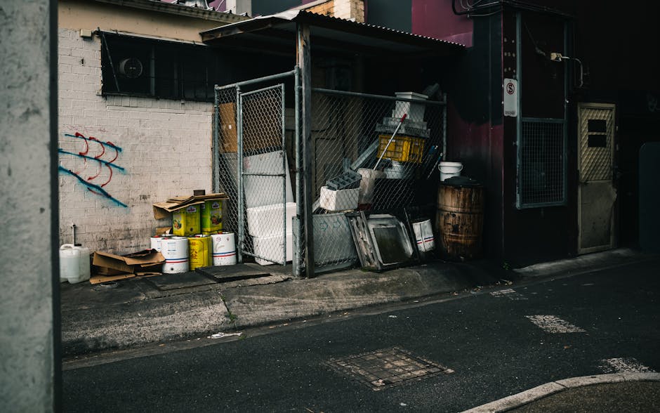 A designated outdoor storage area on a paved urban street shows a collection of various waste items and materials. In the foreground, there are several yellow containers and white buckets stacked on top of each other, alongside flattened cardboard boxes and a large white plastic canister. Behind these, a metal fence surrounds a small structure with a corrugated roof, containing miscellaneous objects including metal trays, plastic storage bins, and a yellow plastic crate. To the right, a weathered wooden barrel and a white plastic bowl are placed next to a dark-colored building with a door and a window, both secured with metal mesh. The scene is lit by natural daylight, with subtle shadows cast on the wall and ground, highlighting the textures of the materials—rough concrete, corrugated metal, and weathered wood. This setup appears to support private waste disposal efforts, aligned with alternative rubbish removal services offered by companies like Rubbish Removal Kingston, rather than official council collection. The overall environment indicates an accessible area for loading waste, consistent with independent rubbish clearance or on-site waste management activities common in urban settings.