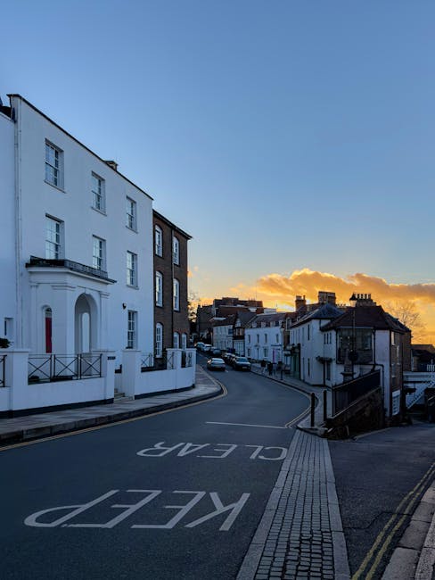 A quiet residential street during sunset in Kingston Hill, featuring a gentle downhill slope with a mix of white and brick buildings lining the pavement. On the left side, a large white multi-story building with flat roof, small balcony, and decorative black railings in front, stands prominently at the street corner. The road is paved with smooth asphalt, displaying the words 'KEEP CLEAR' painted in white, indicating parking restrictions. The curve of the road extends into the distance, where a line of houses with varied architectural styles and dark roofing tiles is visible. The sky is partly cloudy with a warm orange and yellow glow near the horizon, casting a soft light over the scene. The environment appears calm and well-maintained, with street lamps and a slight incline leading to lower residential buildings, consistent with private waste collection and on-site clearance services often utilized by independent operators, such as Rubbish Removal Kingston, in suburban areas like Kingston Hill KT2.