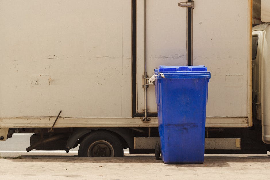 A large blue plastic wheeled bin with a closed lid is positioned on a paved surface beside a white, metal-sided delivery or utility van. The bin appears to be made of durable polycarbonate or similar material with a slightly textured surface and visible smudges or dirt marks on its exterior. It is placed on the right side of the image, leaning slightly against the side of the van, which has a vertical black handle, a metallic hinge, and a lock mechanism near the top. The van's side is plain, with some subtle signs of weathering or dirt. Along the bottom edge of the van, part of the black wheel and suspension system are visible, resting on a concrete or tarmac surface. The background suggests an outdoor environment, possibly a street, driveway, or loading area, where waste collection or rubbish removal activities might take place. The scene is lit evenly with natural light, emphasizing the utilitarian nature of the items and their surroundings, reflecting the type of equipment involved in private waste or bulk rubbish disposal services carried out by Rubbish Removal Kingston.