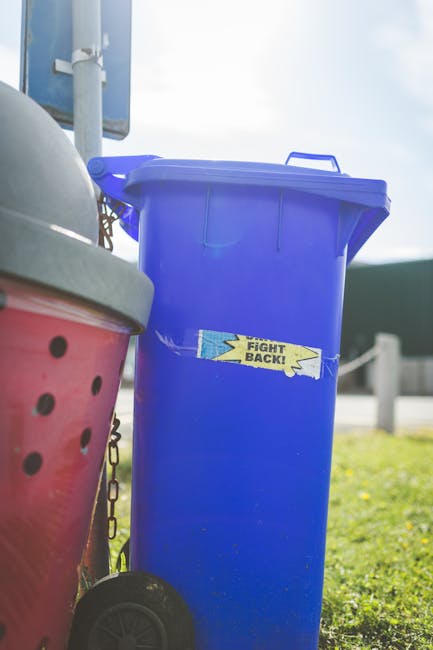 The image shows a row of four large wheeled rubbish bins situated on a paved residential sidewalk in front of a brick building and a parked grey van. The bins are made of black plastic with textured surfaces, and each has a different colored lid: two yellow, one blue, and one black, all with a smooth, matte finish. The bins are positioned side by side, with the yellow-lidded bin on the far left, followed by the blue-lidded bin, and two black-lidded bins on the right, slightly taller. Behind the bins, a residential street scene is visible with an out-of-focus vehicle and suburban houses, suggesting an area where private waste collection services like Rubbish Removal Kingston can provide alternative rubbish disposal options. This setup is typical of private waste handling, particularly where residents prefer on-site or independent collection of bulky waste or general rubbish, aligning with the context of Kingston Council rules for bulky waste kerbside collection. The lighting is natural, with daylight softly illuminating the scene, emphasizing the textures of the bins and the surrounding environment, supporting accessibility by clearly depicting waste management elements relevant to rubbish removal services.