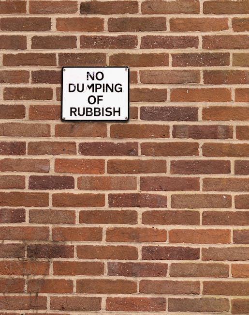 A rectangular metal sign with a white background and black text reading 'NO DUMPING OF RUBBISH' is mounted on a red brick wall. The wall features bricks in varying shades of reddish-brown, laid in a horizontal pattern with light gray mortar joints. The sign is positioned approximately one-third from the top of the image, with bricks extending evenly around it. The surface of the brick wall appears clean and well-maintained, providing a clear contrast to the sign that communicates an environmental message. The lighting in the scene is natural and even, illuminating the wall and sign without strong shadows, emphasizing the textures of the bricks and the smooth surface of the sign. The image captures a typical outdoor setting where waste management, such as rubbish removal or private disposal, might be relevant, and the sign acts as a visual deterrent to illegal dumping, aligning with the broader context of effective rubbish control implemented by local authorities or private waste removal services like Rubbish Removal Kingston.