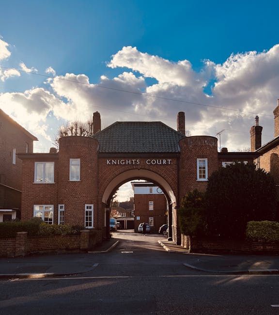 A brick archway entrance labeled 'Knights Court' situated in a residential area with surrounding brick buildings and trees. The archway leads into a small courtyard with parked cars visible in the background. The sky overhead is partly cloudy, with sunlight casting shadows on the buildings and street. The scene is outdoors on a paved road with sidewalks, and there are no visible rubbish or waste items. The image reflects an urban or suburban setting where possible private waste removal or on-site clearance might be conducted by services such as Rubbish Removal Kingston for property maintenance or clearance activities.