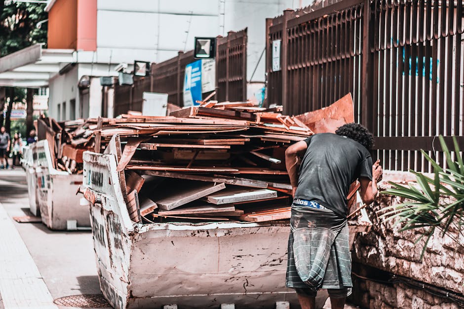 A man with curly dark hair wearing a black t-shirt and gray plaid shorts is standing on a sidewalk next to a large, white, industrial skip filled with broken wooden panels, planks, and construction debris. The wooden materials are in various shades of brown, some with rough, splintered textures, and appear weathered or reclaimed. The skip is positioned adjacent to a stone wall with visible mortar joints on the right side of the image. In the background, there is a tall dark brown metal fence with vertical slats, and beyond that, a glimpse of a modern building with light-colored walls and windows. On the left side, a portion of a busy street scene is visible, with pedestrians walking along a pavement, some partially obscured, and greenery such as trees or bushes can be seen in the distance. The environment is outdoors, illuminated by natural daylight, creating a neutral, daytime atmosphere. This scene exemplifies on-site clearance and the handling of bulky waste materials, relevant to private rubbish removal services like those offered by Rubbish Removal Kingston, supporting alternative waste disposal methods outside of local authority collections.