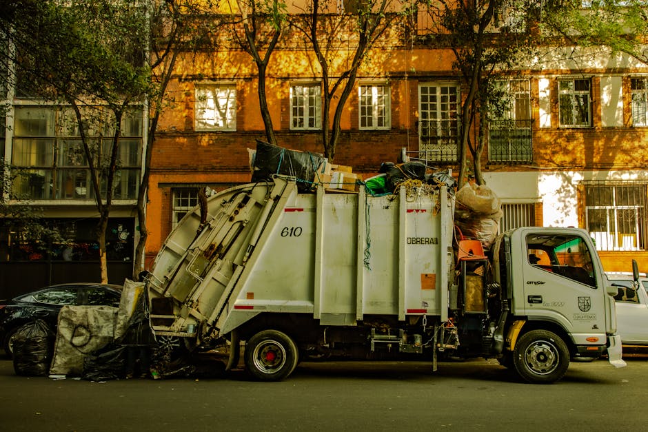 A man with curly dark hair wearing a black t-shirt and gray plaid shorts is standing on a sidewalk next to a large, white, industrial skip filled with broken wooden panels, planks, and construction debris. The wooden materials are in various shades of brown, some with rough, splintered textures, and appear weathered or reclaimed. The skip is positioned adjacent to a stone wall with visible mortar joints on the right side of the image. In the background, there is a tall dark brown metal fence with vertical slats, and beyond that, a glimpse of a modern building with light-colored walls and windows. On the left side, a portion of a busy street scene is visible, with pedestrians walking along a pavement, some partially obscured, and greenery such as trees or bushes can be seen in the distance. The environment is outdoors, illuminated by natural daylight, creating a neutral, daytime atmosphere. This scene exemplifies on-site clearance and the handling of bulky waste materials, relevant to private rubbish removal services like those offered by Rubbish Removal Kingston, supporting alternative waste disposal methods outside of local authority collections.