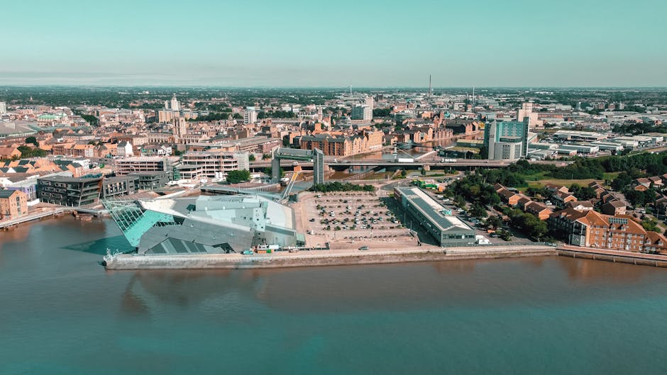 An aerial view of Kingston upon Thames showcasing the Thames River in the foreground, with a modern building featuring angular, silver-grey metallic surfaces and large glass windows situated along the riverbank. To the right of the building, there is a spacious parking lot filled with numerous parked cars. Behind the waterfront, the cityscape extends with a mixture of historical brick buildings and contemporary structures, including office towers and residential blocks. Bridges span across the river in the distance, connecting different parts of the urban area. The scene is illuminated by natural daylight under a clear sky, with the environment appearing vibrant and well-maintained. This image captures a distinctive section of Kingston, highlighting its blend of modern architecture and traditional city elements, relevant to discussions around urban development, infrastructure, and private waste management services like those offered by Rubbish Removal Kingston for efficient rubbish removal and site clearance in the area.