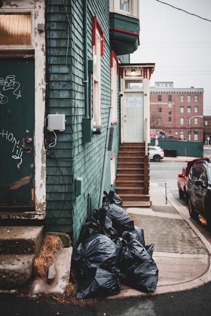 A row of black rubbish bags filled with waste stacked at the base of a green, weathered wooden building with shingle siding. The bags are situated next to a concrete sidewalk and small set of wooden stairs leading up to a white door with a small porch roof. Nearby, a metal electrical box and utility cable are mounted on the building's exterior wall. In the background, there is a parking area with parked cars and a red brick industrial-style building further away under an overcast sky. The scene appears to depict an area awaiting waste collection or disposal as part of private rubbish removal services, with the debris placed neatly outside the building for pickup.