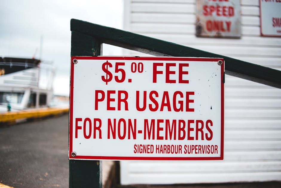 A close-up of a rectangular white sign with bold red lettering, mounted on a black wooden post at a harbor or marina. The sign displays a fee of five pounds for non-members to use the facility, reading '$5.00 FEE PER USAGE FOR NON-MEMBERS', and is signed by the harbour supervisor at the bottom. In the background, parts of boats with white hulls and crewed rigging are visible, along with a paved surface and additional signage partially seen to the right, indicating rules or restrictions. The environment appears overcast, with diffused natural light highlighting the sign and surrounding maritime setting, which relates to private or independent waste handling options often associated with harbour facilities, or alternatively, waste disposal at maritime sites managed by private services such as Rubbish Removal Kingston.