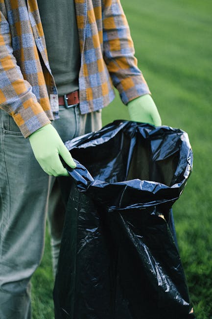 This image shows four large black refuse sacks made of shiny, crinkled plastic material, stacked against a black metal fence on a paved sidewalk. The sacks appear to contain household waste or rubbish, with some of the bags displaying printed text in black ink. Behind the fence, there are dense, leafy bushes and trees, partially illuminated by natural light, indicating an outdoor setting. The scene captures the typical context where private waste collection services, such as those provided by Rubbish Removal Kingston, might be involved in the clearance or removal of waste from residential or commercial sites. The overall tone is neutral and professional, with a focus on accurately describing the visible objects and surroundings, supporting accessible understanding and SEO relevance for rubbish removal services in Kingston.
