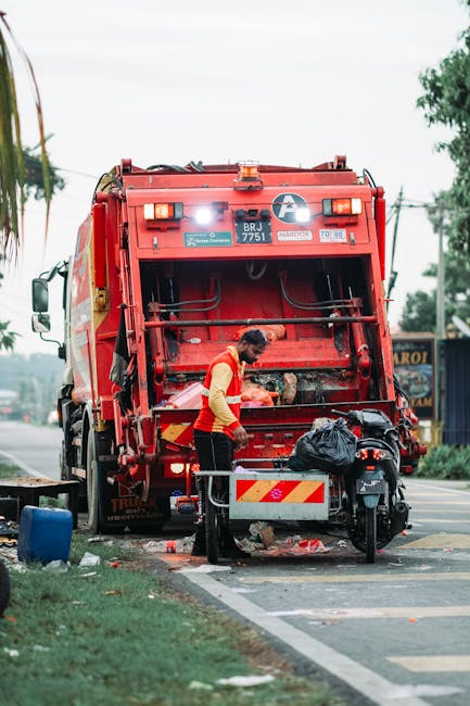 A worker wearing a high-visibility vest and dark trousers is operating a red rubbish collection truck on a street. The truck's rear hopper is open, revealing mechanical components and a bright white light illuminating the area. The worker is standing close to the truck, managing black refuse bags that are placed near a small metal barrier. The scene takes place on a paved road with a grassy verge and a sidewalk, and there are trees and buildings in the background, indicating an urban or suburban environment. The truck appears to be in the process of collecting waste, consistent with services provided by Rubbish Removal Kingston for on-site or private rubbish disposal. The overall scene is lit naturally, likely during daylight hours, and the truck is positioned near a side street or parking area to facilitate rubbish collection or alternative waste handling in a professional waste management context.