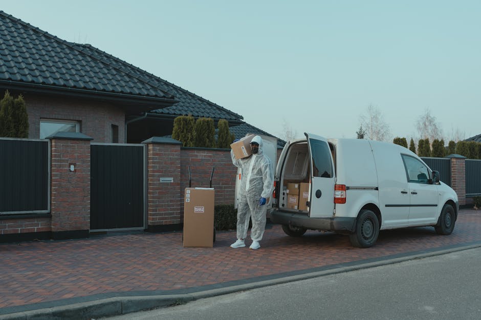 A person dressed in full protective work gear, including a white coverall, gloves, and a face mask, is standing next to an open white delivery van parked on a paved driveway in front of a residential property. The individual is holding a cardboard box, with one hand carrying it on their shoulder while the other hand rests near a stack of boxes inside the van, which contains several packages. Adjacent to the person is a cardboard waste bin with a label, placed on the brick-paved surface. The background reveals a modern house with dark grey tiled roofing, brick walls, and a driveway bordered by a black metal and brick fence. Tall, neatly trimmed shrubs and trees are visible behind the fence, and the scene is illuminated by natural daylight, suggesting a typical residential street. This setup indicates a professional waste clearance or rubbish removal process, possibly conducted by a service like Rubbish Removal Kingston, handling household debris efficiently in an organized manner.