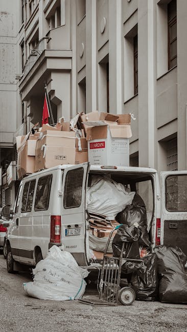A white commercial van parked on a city street with its rear doors open, revealing an interior filled with various types of waste. The load includes large black plastic garbage bags, flattened cardboard boxes, and miscellaneous debris, some stacked on a small trolley positioned at the back of the van. Several flattened cardboard boxes, some with visible printed labels, are placed on the roof of the vehicle, indicating a collection of accumulated rubbish awaiting removal. The van is situated in front of a multi-story building with a grey, stone facade featuring rectangular windows and decorative architectural elements. The scene is set during daylight hours with natural lighting, capturing an environment typical of private waste collection or rubbish removal services in an urban area. The presence of local waste removal suggests an alternative to municipal disposal, aligning with independent rubbish collection options provided by companies like Rubbish Removal Kingston.