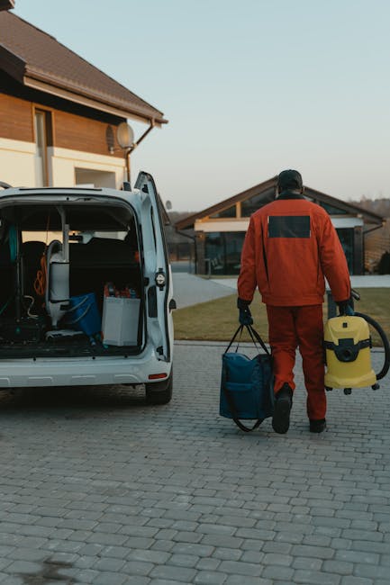 A person dressed in full protective work gear, including a white coverall, gloves, and a face mask, is standing next to an open white delivery van parked on a paved driveway in front of a residential property. The individual is holding a cardboard box, with one hand carrying it on their shoulder while the other hand rests near a stack of boxes inside the van, which contains several packages. Adjacent to the person is a cardboard waste bin with a label, placed on the brick-paved surface. The background reveals a modern house with dark grey tiled roofing, brick walls, and a driveway bordered by a black metal and brick fence. Tall, neatly trimmed shrubs and trees are visible behind the fence, and the scene is illuminated by natural daylight, suggesting a typical residential street. This setup indicates a professional waste clearance or rubbish removal process, possibly conducted by a service like Rubbish Removal Kingston, handling household debris efficiently in an organized manner.